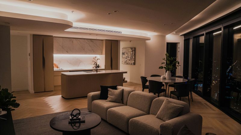 An open-plan living and dining area with a beige sectional sofa, round coffee table, kitchen island, and warm recessed lighting at night.