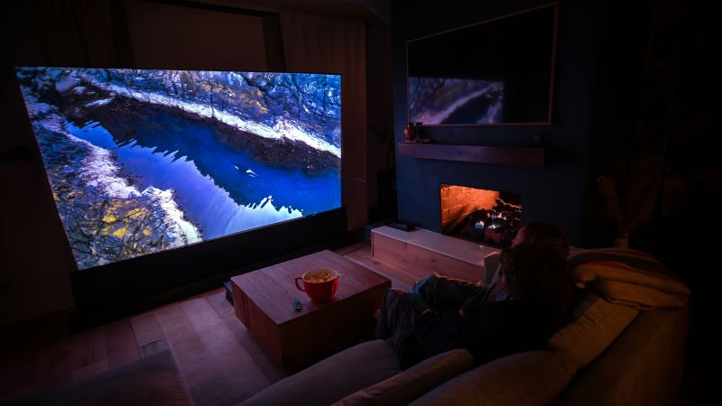 A person sitting on a sofa in a dark living room watching a large wall-mounted screen, with a fireplace glowing nearby.