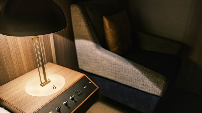 A modern table lamp on a wooden side table beside a gray upholstered armchair, softly lit in a cozy corner.