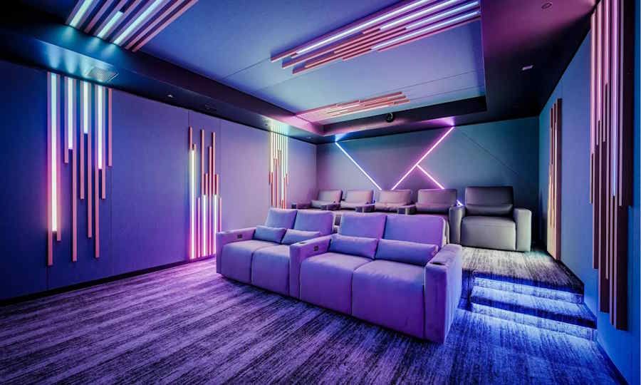 Modern luxury home theater with tiered plush seating and vibrant RGB LED lighting accents on walls, ceiling, and steps, creating a futuristic ambiance with purple, pink, and blue hues.