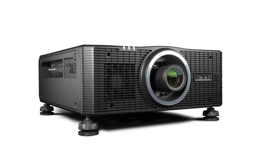 Barco high-performance projector in black, shown at an angle highlighting its prominent lens, ventilation grille, adjustable mounting feet, and side input panel.